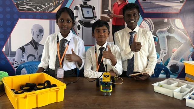 Three students in school uniform give a thumbs up behind the wheeled robot they built